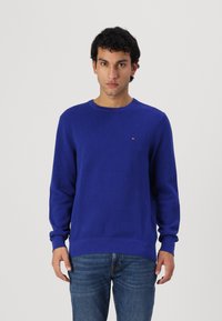 Pull bleu tricoté avec un col rond et des poignets côtelés, arborant un petit logo sur la poitrine. Assorti avec un jean en denim.
