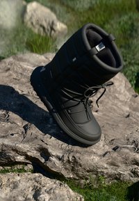 Black insulated winter boot with a padded design, drawstring closure, and textured rubber sole, resting on a rocky surface.