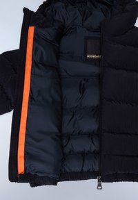 Svart isolert pufferjakke innvendig med oransje detaljer langs glidelåsen, quiltet tekstur, og et svart stoffmerke på brystlommen.