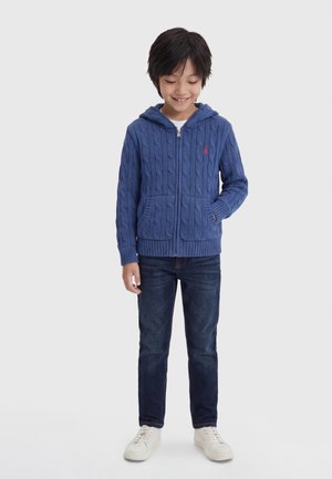 Young boy wearing a blue cable-knit zip-up hoodie, dark jeans, and white sneakers, standing with one hand in pocket, smiling and looking down.