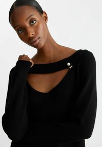 Maglione nero in maglia con un ampio scollo a spalle scoperte, caratterizzato da un dettaglio di spilla con perle e fiocco sulla spalla sinistra. Texture morbida.