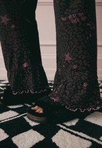 Pantaloni a zampa di elefante con stampa leopardata nera, decorati con testi rosa e motivi a stella, abbinati a ciabatte nere pelose con fascia arricciata e suola in sughero.