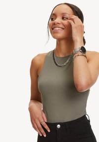 Olijfgroene mouwloze bodysuit met een hoge halslijn. Model is voorzien van gelaagde zilveren kettingen en een zilveren horloge. Zwarte high-waisted broek.