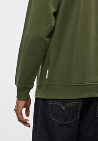 Oliviroheline fliisriie, millel on tekstuuriga viimistlus, soonilised varrukaotsad ja väike kaubamärgi silt ääres. Sobitatud tumedate teksadega, millel on kollane õmblus.