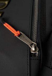 Schwarzer Stoff mit einem metallischen Reißverschluss und einem silbernen Ziehband, das mit "SUPERDRY" beschriftet ist, sowie einem orangefarbenen Kordelzug. Die Nähte ergänzen das Design.
