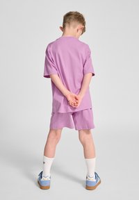 Lila Baumwoll-T-Shirt und Shorts, beide locker sitzend. Das Kind trägt weiße Knöchelsocken und hellblaue Schuhe mit braunen Sohlen.