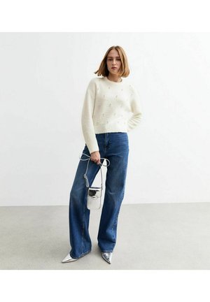 Suéter blanco de punto con detalles texturizados, combinado con jeans de tiro alto azul. Zapatos plateados de punta y un pequeño bolso metálico completan el look.