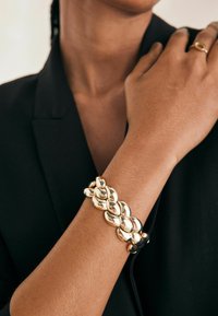 Bracelet en métal doré présentant des formes ovales interconnectées avec une surface brillante et réfléchissante. Porté au poignet, contrastant avec un vêtement noir.