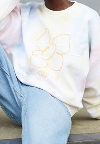 Persoon draagt pastel tie-dye sweatshirt met gele bloemprint en lichtblauwe spijkerbroek, zittend met één been gebogen.