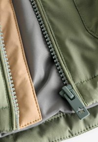 Intérieur d'une veste en tissu vert, avec une doublure beige et grise, fermeture éclair exposée avec un détail en forme d'étoile, mettant en valeur une texture lisse et des coutures discrètes.