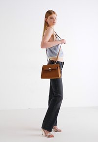 Brun læderhandtaske med en struktureret design, der har en klaplukning, topgreb og justerbar strop, parret med mørke jeans og en stribet tanktop.