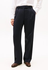 Marineblauwe broek met een getailleerde pasvorm, voorzien van plooien, riemlussen en zijzakken, gecombineerd met bruine loafers, op een effen witte achtergrond.