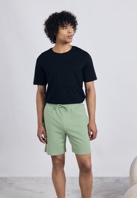 Tricou negru cu mânecă scurtă asociat cu pantaloni scurți verzi deschis, prevăzuți cu șnur. Ambele articole sunt realizate dintr-un material moale și respirabil.