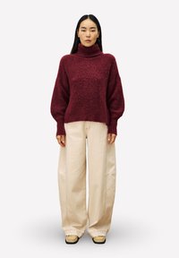 Frau in einem burgunderfarbenen Rollkragenpullover, weiten beigen Hosen und beigen Schuhen mit quadrater Spitze, die vor einem schlichten hellen Hintergrund steht.