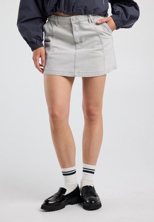 Persoon draagt een lichtgrijze denim minirok, witte sokken met zwarte strepen, zwarte loafers en een donkere lange mouwen jas met handen gedeeltelijk in de zakken.