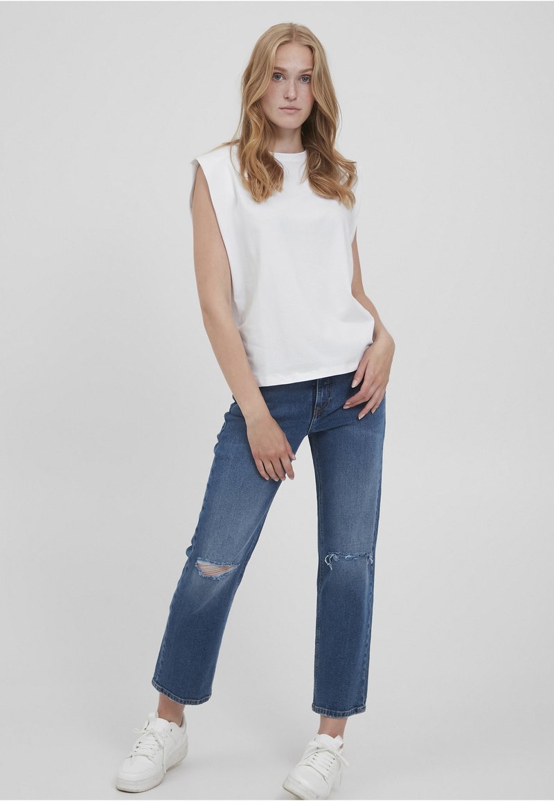 Hvid ærmeløs top med en rund halsudskæring, paired med slidte blå jeans og hvide sneakers. Casual outfit mod en lys grå baggrund.