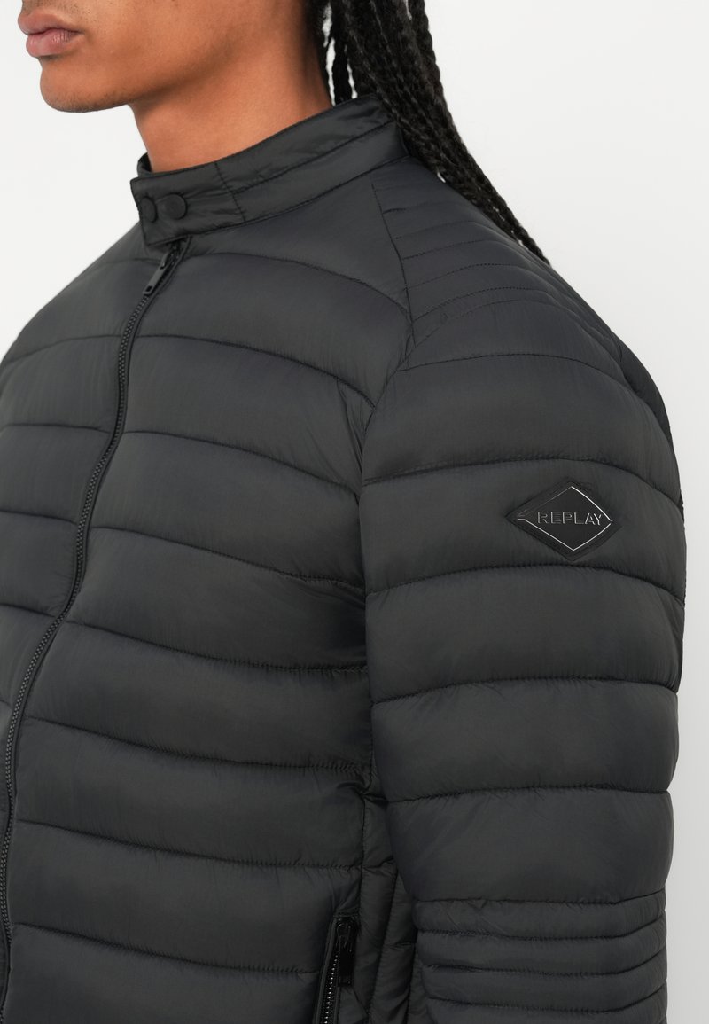Doudoune noire matelassée avec col montant, comportant une fermeture éclair à l'avant et des poches latérales. Inclut un logo en forme de diamant sur la manche gauche.
