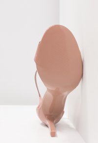 Sandale à talons hauts en cuir verni rose avec un design ouvert à l'avant, une fine lanière et une semelle texturée. L'angle montre le talon et le placement de la base.