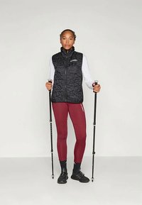 Μαύρο γιλέκο με γεωμετρικό ανάγλυφο σχέδιο, φορεμένο πάνω από ένα λευκό μπλουζάκι με μακριά μανίκια. Το μοντέλο φοράει επίσης βυσσινί κολάν και κρατάει μπαστούνια πεζοπορίας.