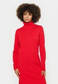 Abito rosso con collo alto realizzato in morbido e elastico tessuto a maglia. Maniche lunghe, polsini a costine e una fessura laterale per facilitare i movimenti.