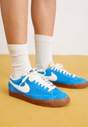 Persoon draagt witte geribbelde enkelsokken en blauwe Nike-sneakers met witte Swoosh en bruine zolen, staand op een gele ondergrond.