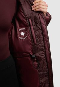 Burgundska prešita jakna z gladkim podlogom, ki ima oznako logotipa in nastavljivo vrvico znotraj. Žep na zadrgo na levi strani.