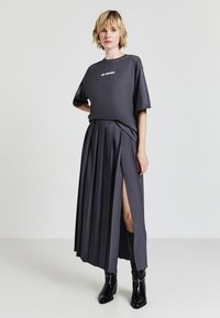 Donna con capelli biondi corti che indossa una maglietta oversize grigia scura con il testo "NO DRAMA", una gonna plissettata con uno spacco sulla coscia e stivali neri.