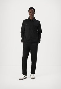 Casaco casual preto com dois bolsos no peito, fecho de botão, combinado com calças pretas a condizer e sapatos brancos com riscas pretas.