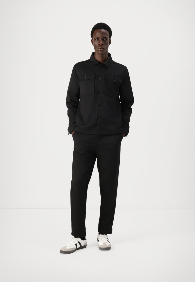 Casaco casual preto com dois bolsos no peito, fecho de botão, combinado com calças pretas a condizer e sapatos brancos com riscas pretas.