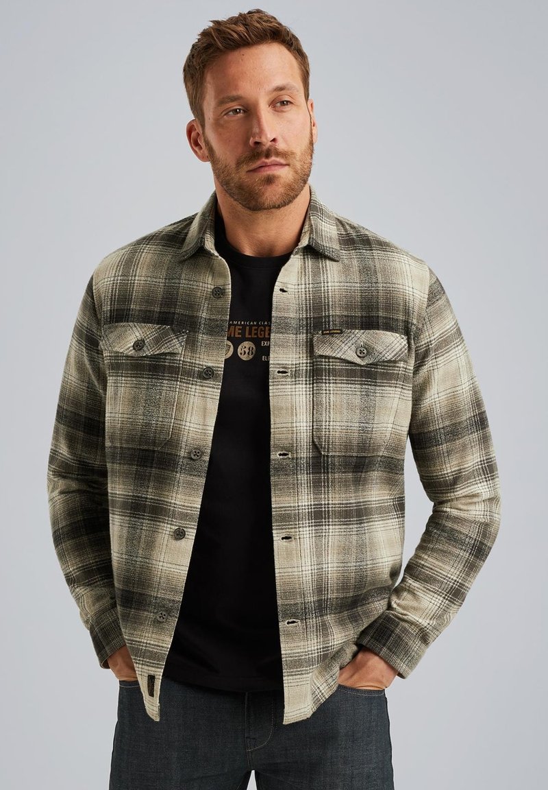 Rutig flanellskjorta i beige och svarta toner med knappstängning framtill, två bröstfickor och krage. Bärs över en svart grafisk t-shirt.
