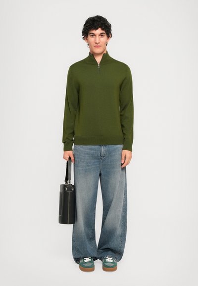 Ung mann står, iført grønn zip-up genser, vide falmede jeans, grønne sneakers, holder en svart lærveske på siden.