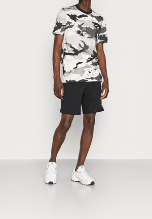 Chemise à manches courtes à motif camouflage en gris et noir, associée à un short noir. Des chaussures de sport blanches avec des accents noirs complètent la tenue.