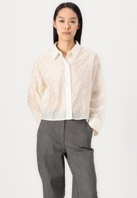 White embroidered blouse with floral patterns, button closure, and collar. Cropped design, lightweight fabric, worn with gray striped trousers.