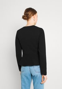 Frau mit braunem Haar im Dutt, die silberne Creolen trägt, ein enges schwarzes Langarmoberteil und hellblaue hochgeschnittene Jeans anhat, steht vor einem weißen Hintergrund.