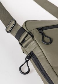 Olijfgroene schoudertas van stevig stof, met een zwarte rits, een verstelbare schouderband en een ronde trekknop. Textuur oppervlakte.