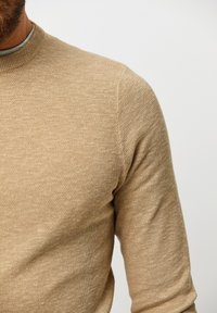 Beige stickad tröja med en strukturerad väv, rund halsringning och långa ärmar, som visar en ljus och mjuk tygkänsla.