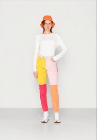 Calças de ganga em patchwork colorido nas cores amarelo, rosa e laranja, combinadas com uma blusa de manga longa branca e um chapéu laranja tipo balde. Inclui ténis brancos.