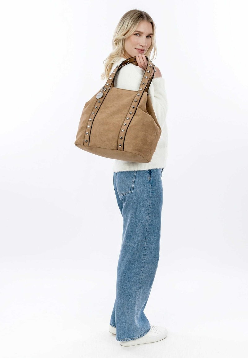 Sac fourre-tout en suède marron avec des accents cloutés et des poignées sombres contrastantes, présentant un corps spacieux et une base arrondie pour la structure.