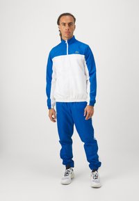 Zils un balts sporta uzvalks ar jaku ar rāvējslēdzēju un stāvošu apkakli, apvienots ar atbilstošām biksēm un baltajiem tenisa apaviem.