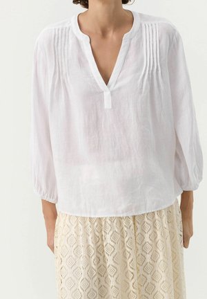 Woman wearing a white long-sleeve blouse with pintuck details and a beige lace patterned skirt.