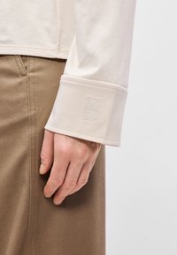 Κρέμα μπλούζα με μακριά μανίκια και μανσέτα που διαθέτει κεν�τημένο λογότυπο. Λείος βαμβακερός υλικός συνδυασμένος με ανοιχτό καφέ παντελόνι από κορίνα.