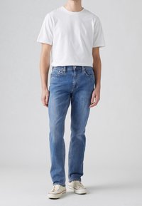 Homme portant un t-shirt blanc à manches courtes, un jean bleu droit et des baskets basses blanches, debout sur un fond uni.
