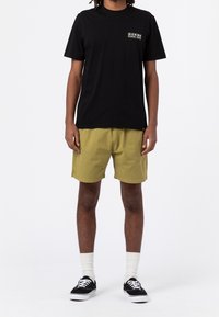 Osoba v černém tričku Dickies, olivově zelených šortkách, bílých ponožkách a černých teniskách stojící na pozadí s jednoduchou bílou barvou.
