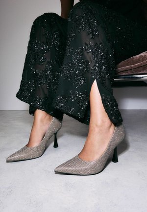 Bisa perak yang berkilau dengan desain ujung runcing, dipadukan dengan celana hiasan sequin hitam yang memiliki belahan samping.