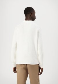 Biały, teksturowany sweter z okrągłym dekoltem i długimi rękawami, w połączeniu z beżowymi spodniami. Miękka tkanina, swobodny krój i minimalistyczny design.