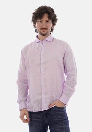 Uomo con capelli ricci che indossa una camicia viola chiaro con bottoni e jeans scuri, in piedi con una mano in tasca su sfondo bianco.