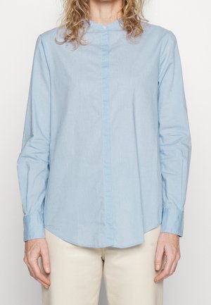 Persona che indossa una camicia azzurro chiaro a maniche lunghe e pantaloni beige, in piedi davanti a uno sfondo chiaro e uniforme con le braccia rilassate lungo i fianchi.
