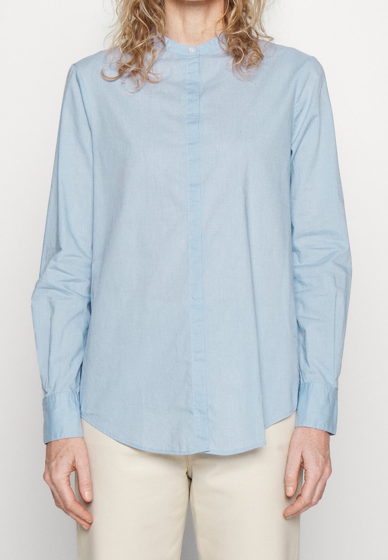 Personne portant une chemise bleu clair à manches longues et un pantalon beige, se tenant devant un fond clair uni, les bras relâchés le long du corps.