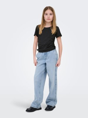 Ung pige stående iført en sort rynket kortærmet top, løse lyseblå jeans og sorte sko mod en ensfarvet hvid baggrund.
