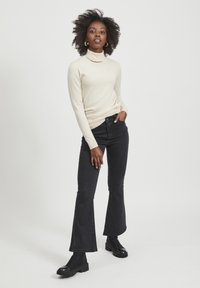 Cremefarvet turtleneck sweater med lange ærmer, parret med sorte flared jeans. Modellen bærer sorte ankelstøvler med en glat tekstur og blank finish.